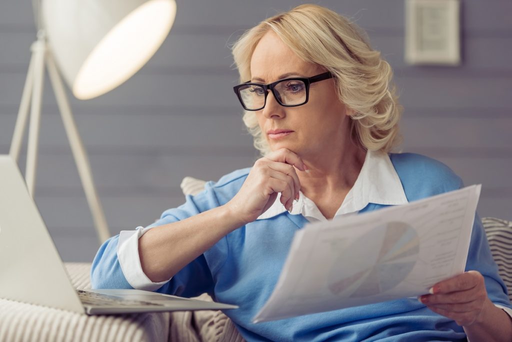 Beautiful old woman in casual clothes and glasses is holding papers and looking at laptop while working at home