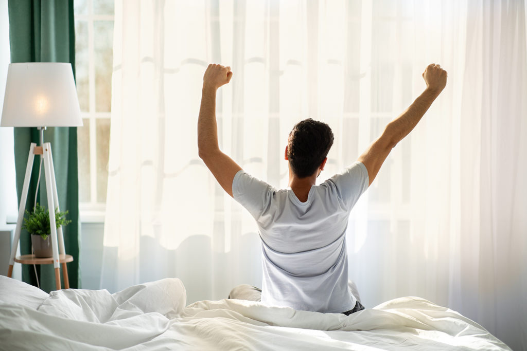 Man sits on the edge of his bed after waking up and stretches his arms towards the ceiling