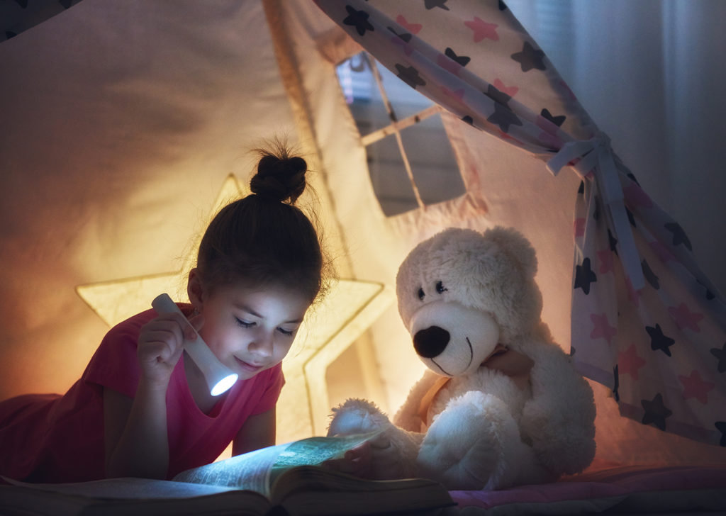 Cute little child is reading a book with flashlights in tent